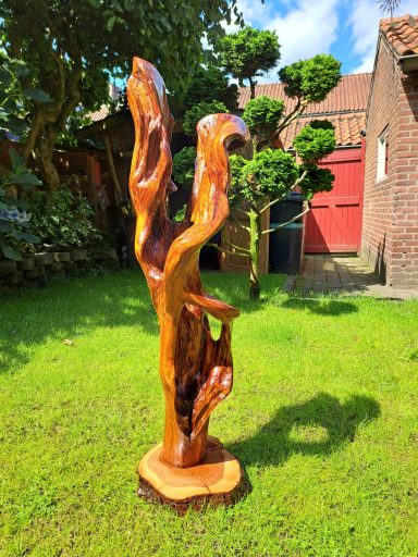 Houten sculptuur met organische vormen, geplaatst in een groene tuin.